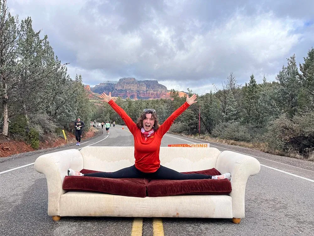 A woman in a red long-sleeved shirt and black leggings performs a split on a couch in the middle of a road, with other runners and scenic red rock mountains in the background.