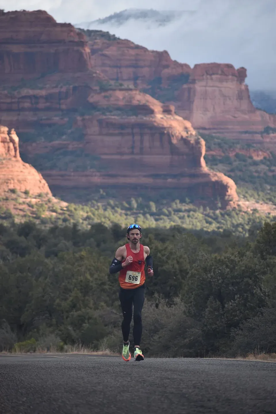 A male runner with bib number 696 runs on a road with dramatic red rock formations and green trees in the background.