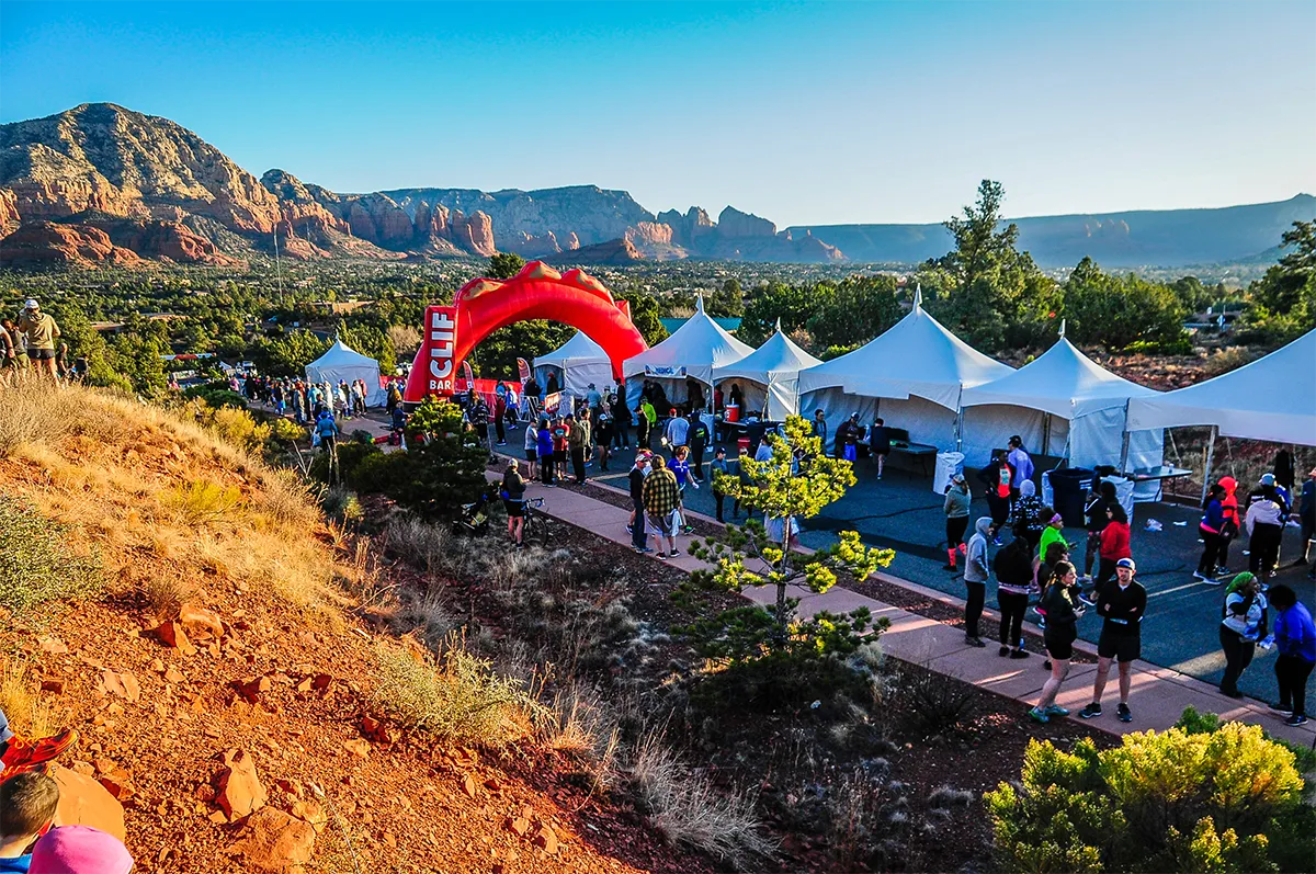 Runners gather near a red inflatable arch at the start of a race, with tents set up and scenic red rock mountains in the background.