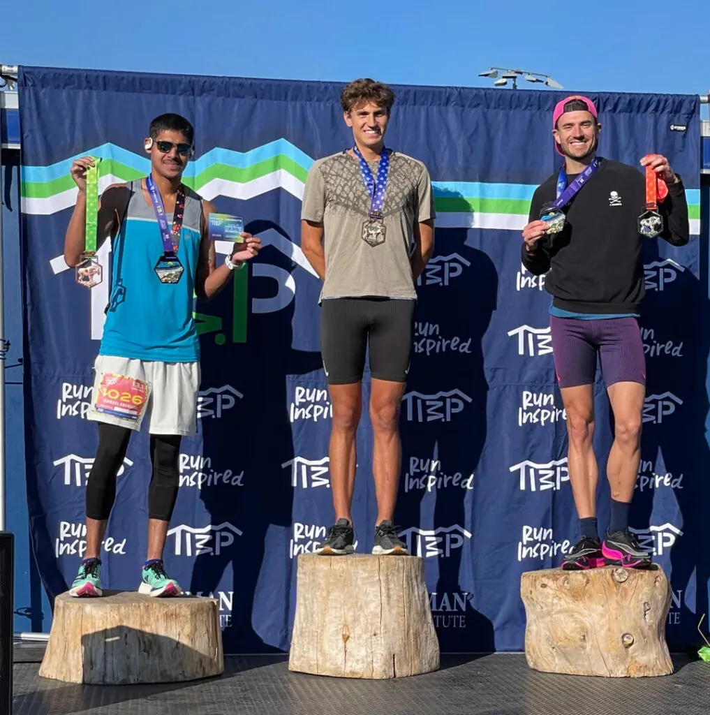 Three male runners on a podium, holding medals and smiling.