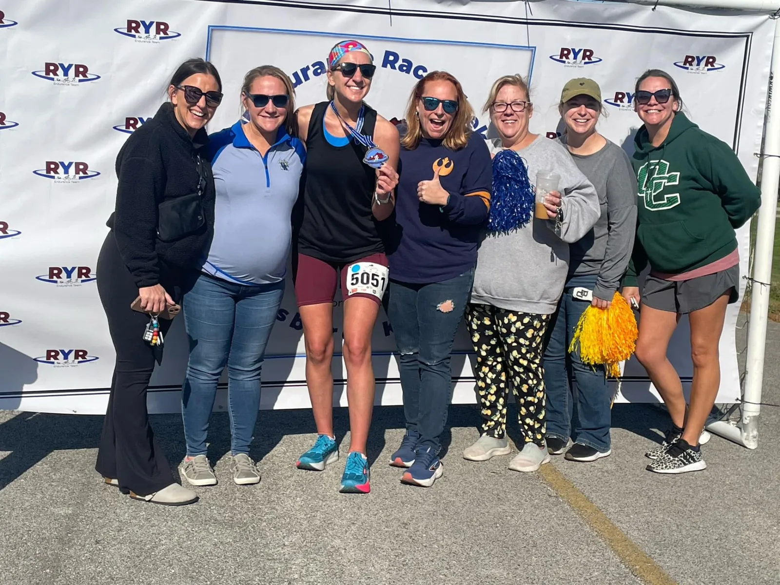 A group of seven women smiling and posing for a photo after a race, with one woman holding up a medal.