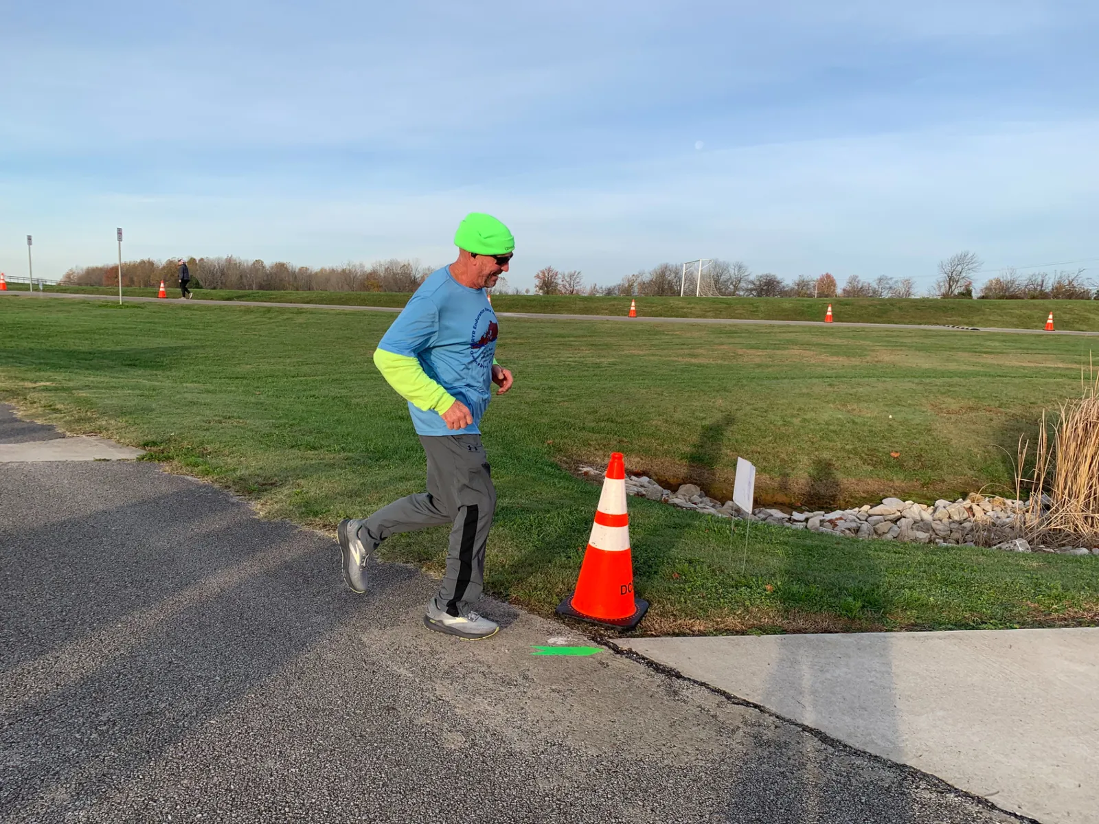 A male runner wearing a blue long-sleeved shirt, bright green beanie, and sunglasses runs on a paved path next to a grassy area with orange traffic cones.