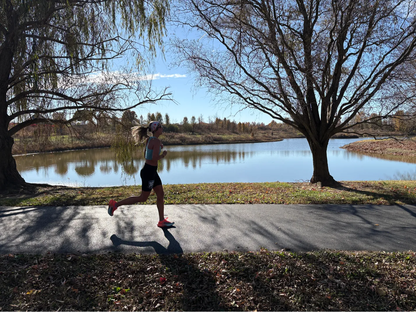 A female runner in profile on a paved path next to a lake, with trees and a clear blue sky.