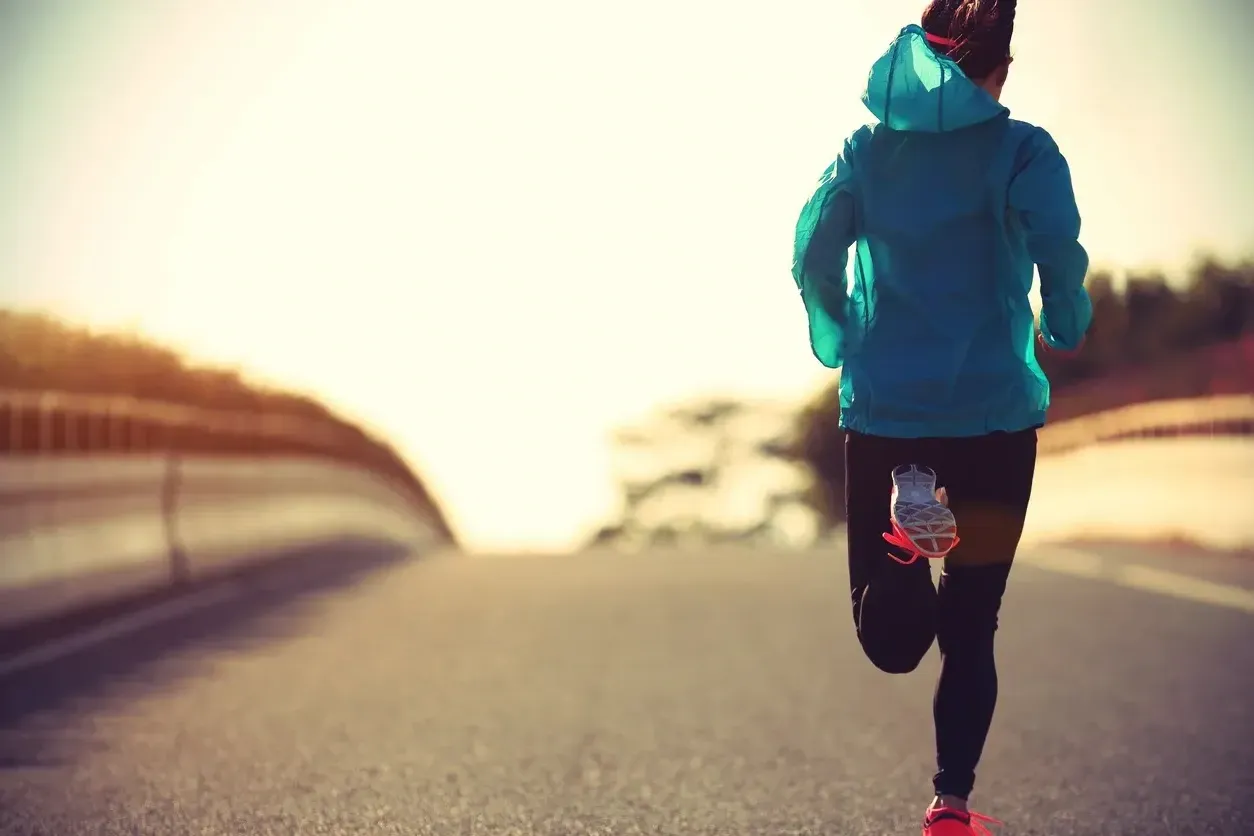 A runner in a blue hooded jacket runs on a road with the sun in the background.