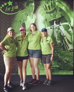 Four women in green shirts and shorts stand in front of a green graphic featuring runners and cyclists.