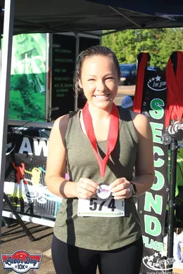 A smiling woman wearing a race medal and bib stands in front of a tent.