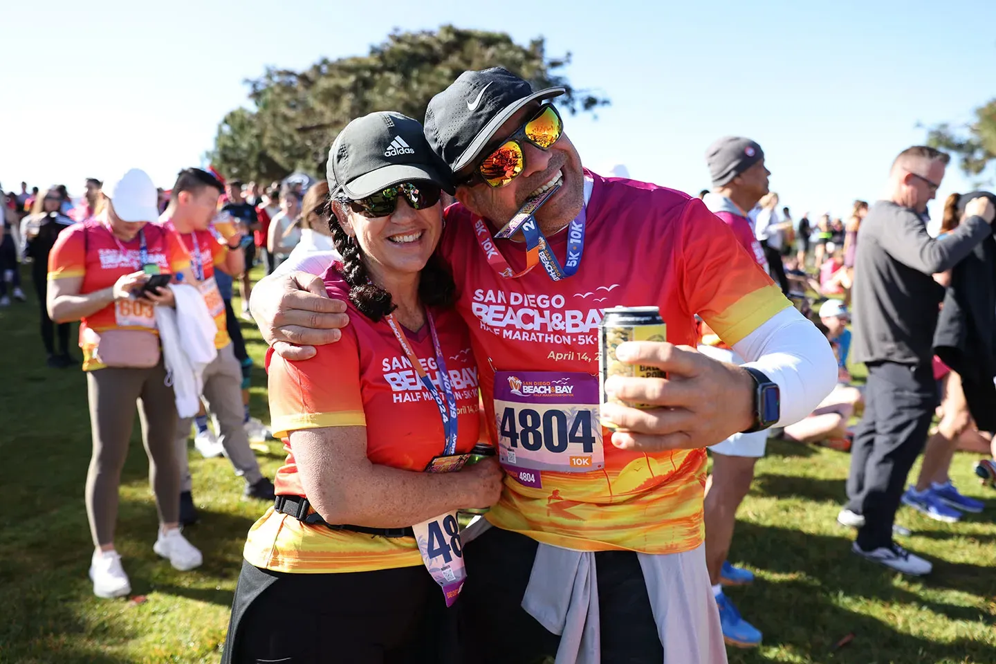 A smiling couple, wearing San Diego Beach & Bay Half Marathon shirts and medals, embrace while holding a can and bib number 4804.