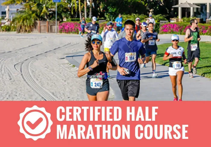 Runners on a paved path next to a sandy beach, with palm trees and buildings in the background. A red banner at the bottom reads "CERTIFIED HALF MARATHON COURSE".