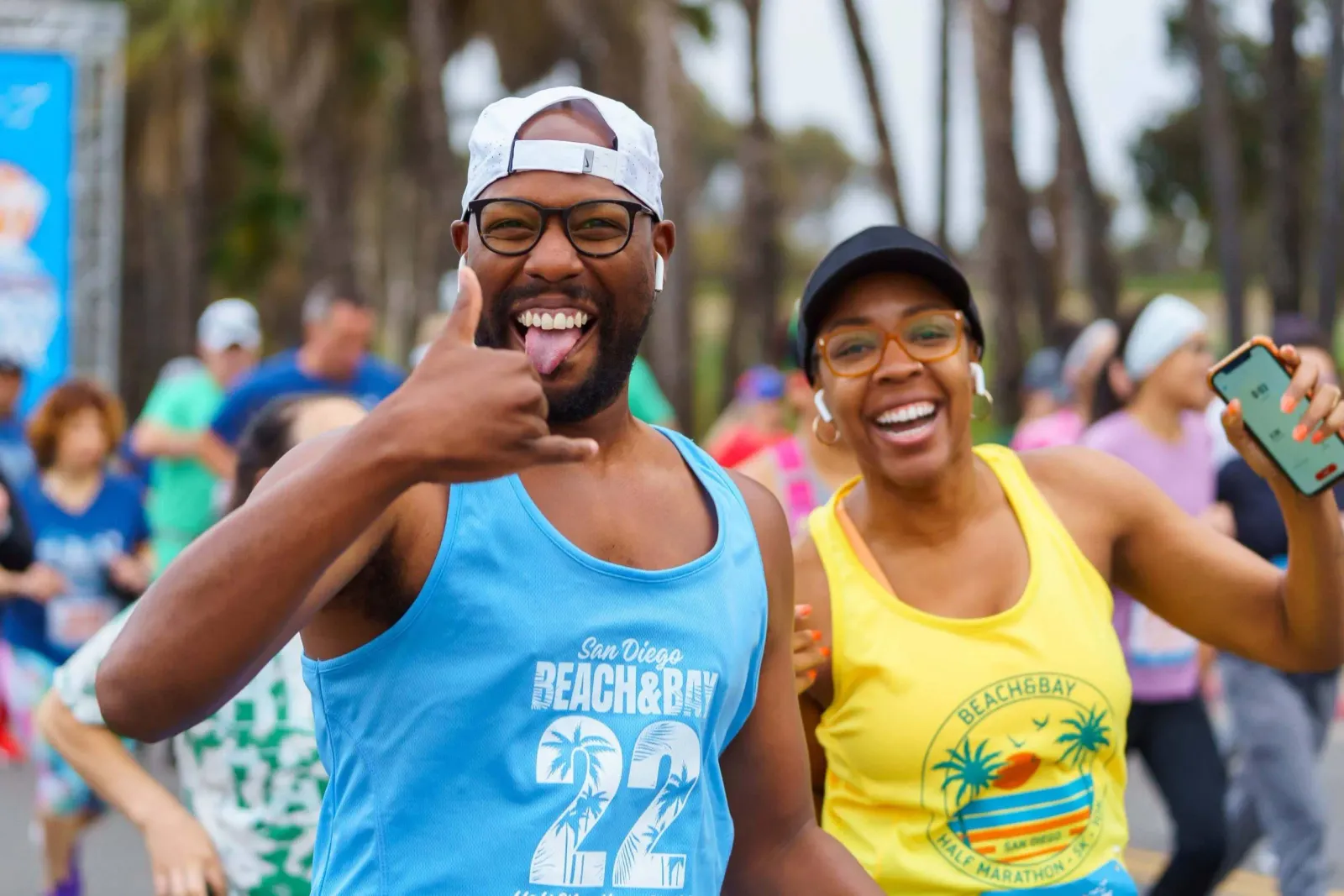Two smiling runners, one giving a shaka sign and sticking out his tongue, and another holding up her phone, participate in the San Diego Beach & Bay Half Marathon.