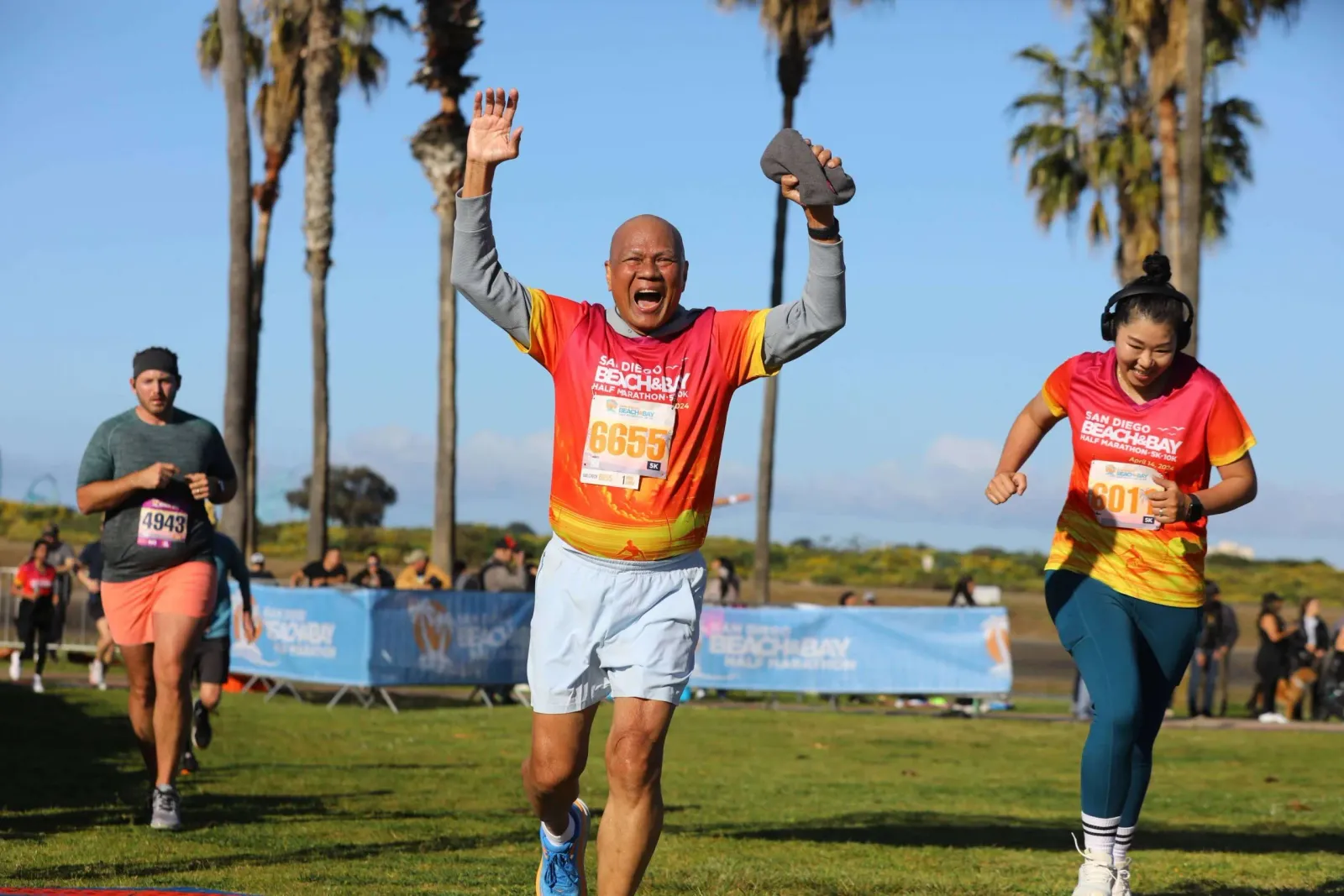 A runner with arms raised in celebration crosses the finish line, with other runners and palm trees in the background.