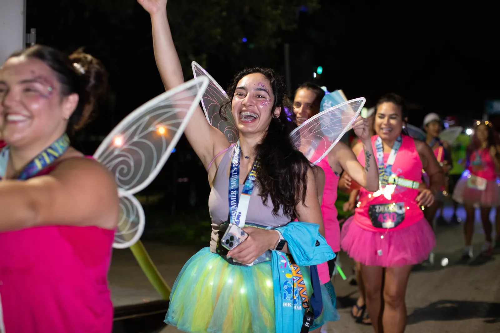 A group of women in fairy costumes, some wearing wings and tutus, smiling and celebrating during a night race.
