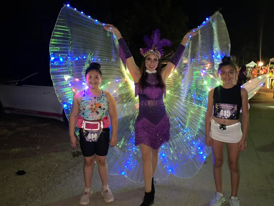 Two young girls and a woman in a purple costume with illuminated wings pose for a photo at a night race.