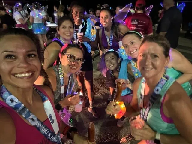 A group of smiling women wearing race medals and some with fairy wings, posing for a photo after a race.