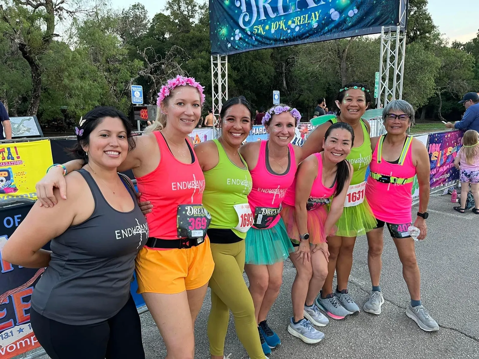 A group of seven women in colorful running attire, smiling and posing for a photo at a race event, with a race banner in the background.