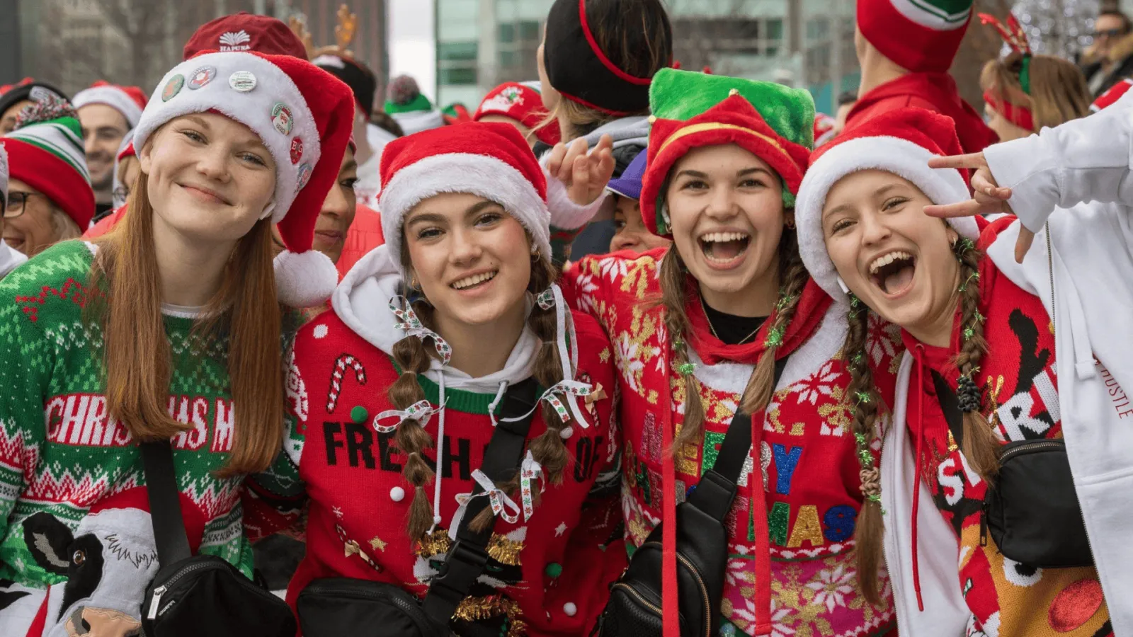 A group of young women wearing Santa hats and festive Christmas sweaters, smiling and laughing at a race event.