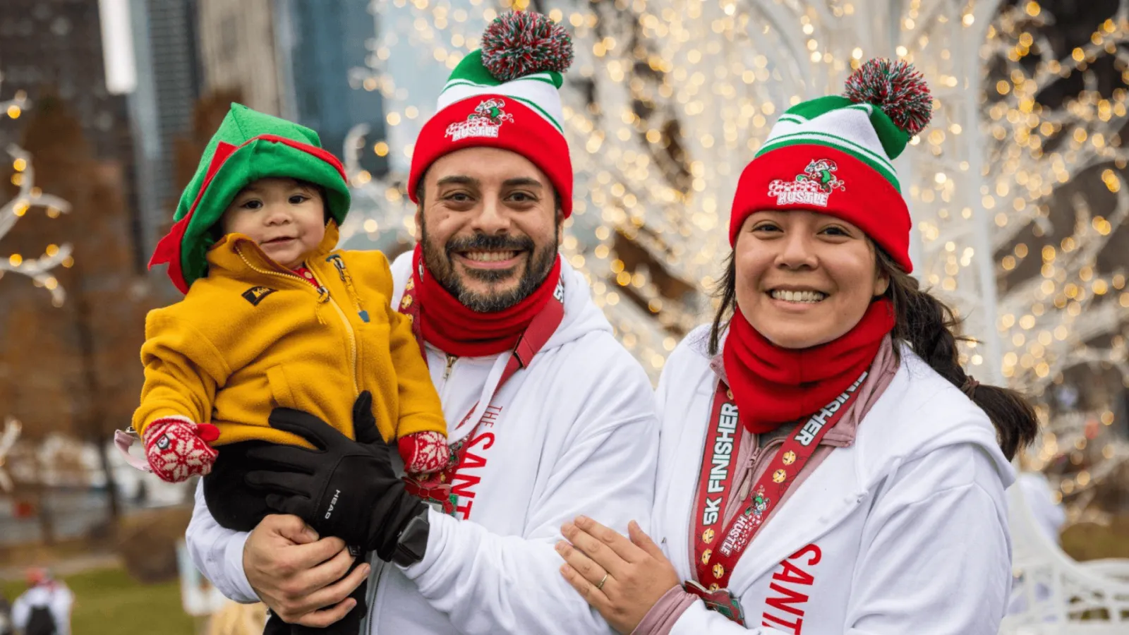 A family, including a baby dressed as an elf, smiles at the camera while wearing Santa hats and race medals, with festive lights in the background.