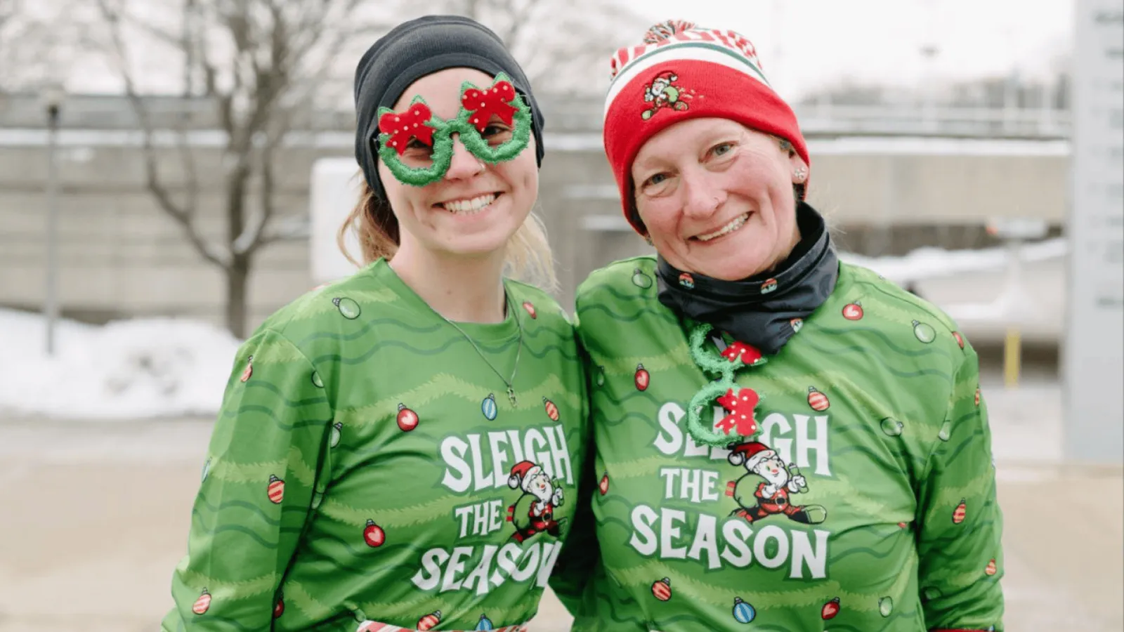 Two smiling women in festive green long-sleeve shirts with Christmas-themed graphics, wearing holiday-themed glasses and hats, posing for a photo at a race.