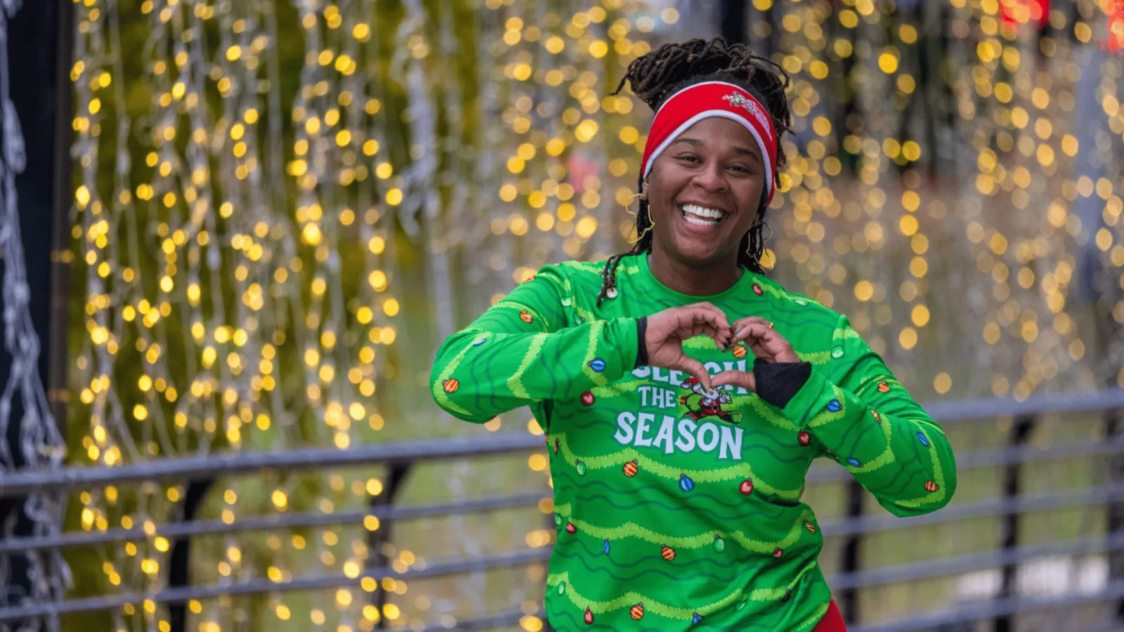 A smiling woman wearing a festive green long-sleeved shirt with Christmas lights and ornaments, and a red headband, forms a heart shape with her hands in front of a background of blurred yellow lights.