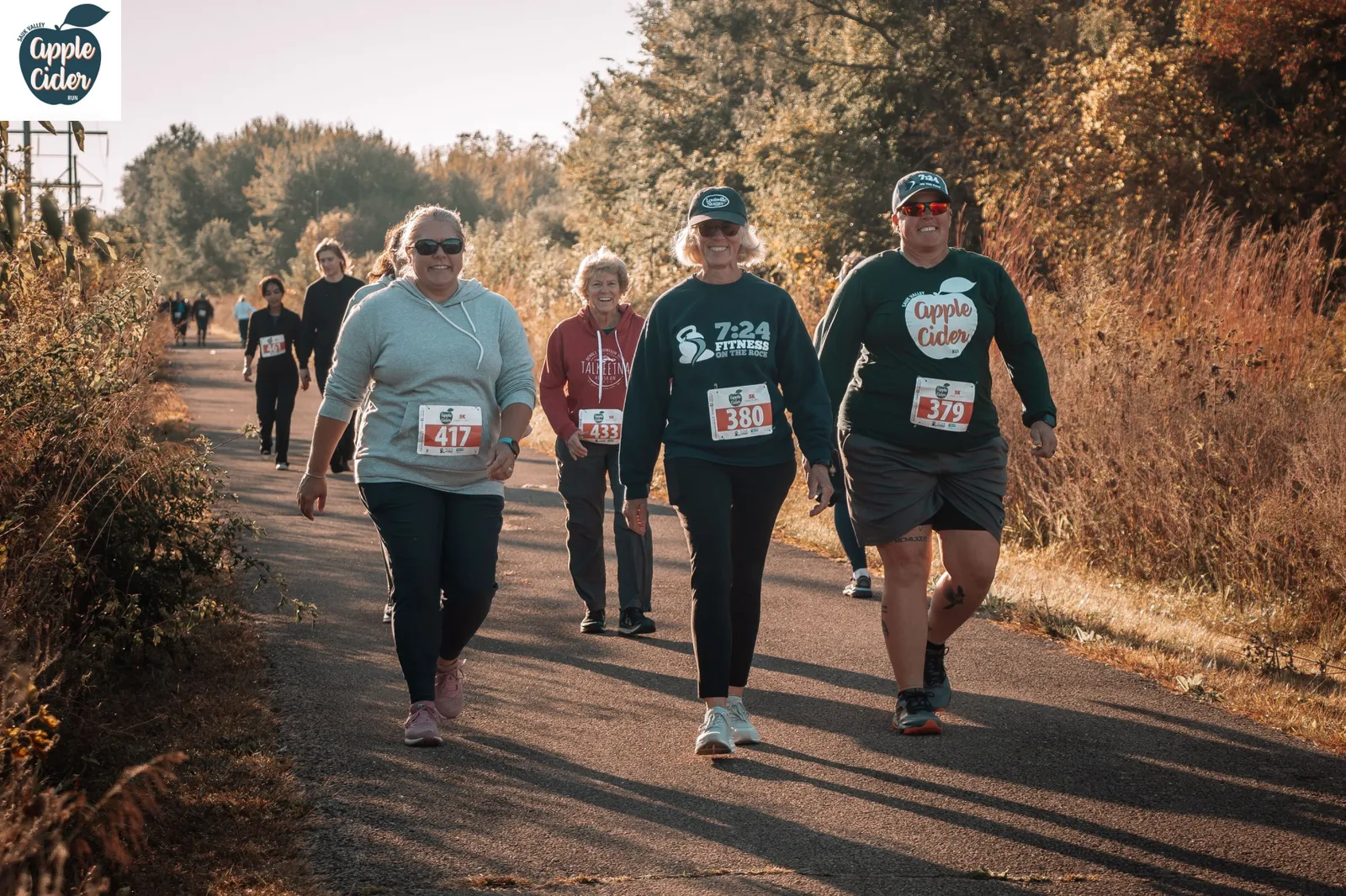 Runners participating in the Sauk Valley Apple Cider Run, with trees and foliage in the background.