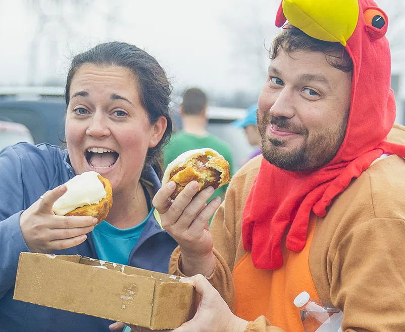 A woman with an excited expression and a man in a chicken costume both holding and eating food items, with other people blurred in the background.