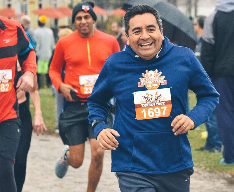A smiling runner wearing a blue hoodie with a 'Turkey Trot' logo and bib number 1697 runs towards the camera.