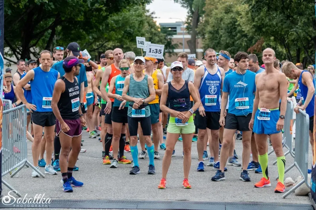 Runners lined up at the start of a race, with bib numbers visible and some holding signs indicating pace groups.