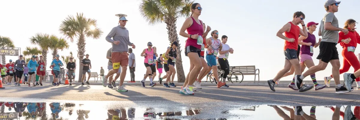 A group of runners participating in a race, with palm trees and a waterfront in the background, under a bright sky.