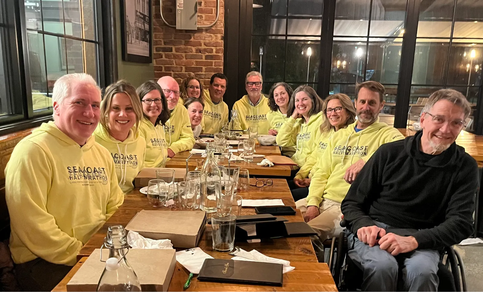 A group of people wearing yellow Seacoast Half Marathon hoodies are seated at a table, smiling and appearing to be celebrating.