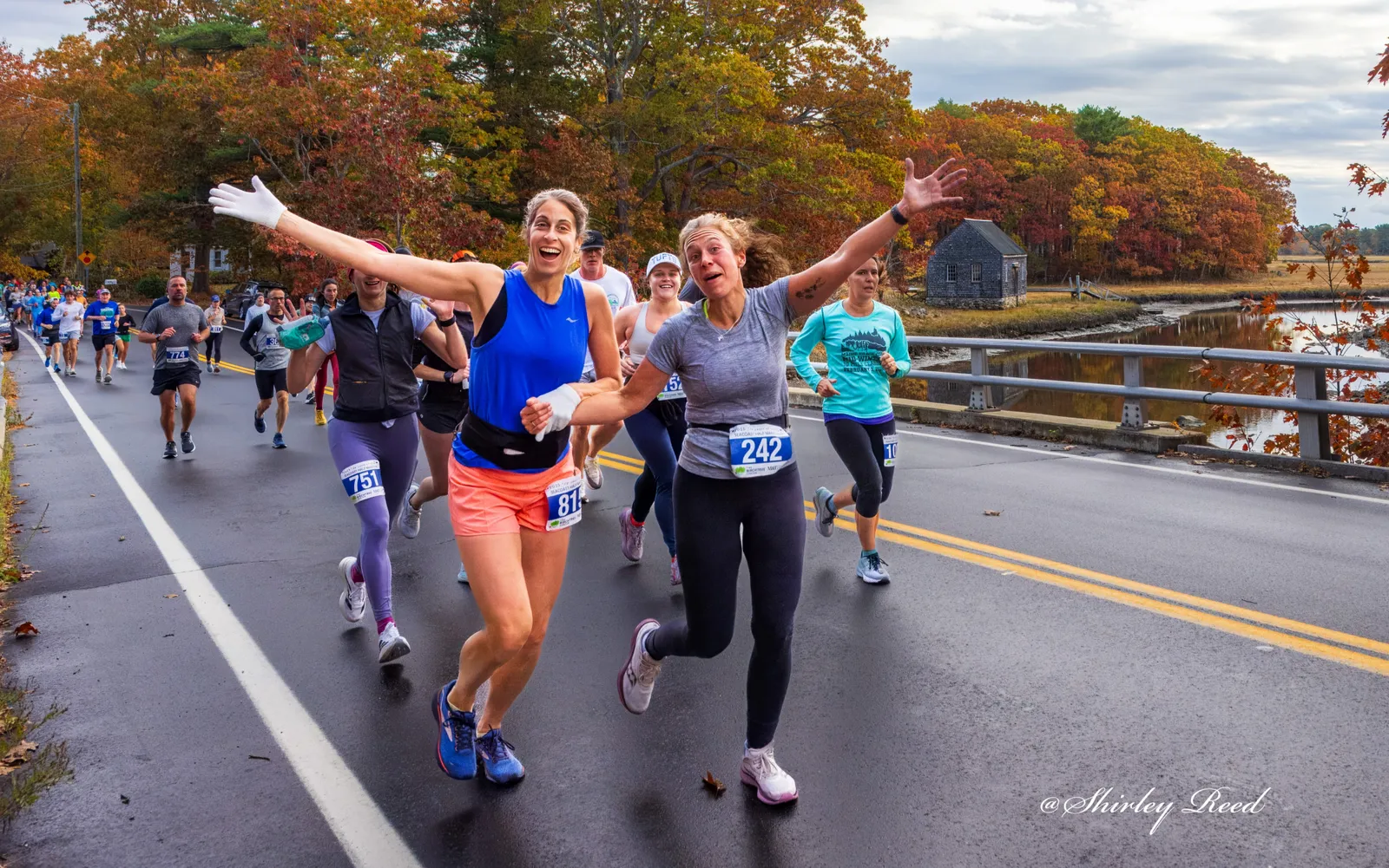 Two female runners with arms outstretched in celebration during a race, with autumn foliage in the background.