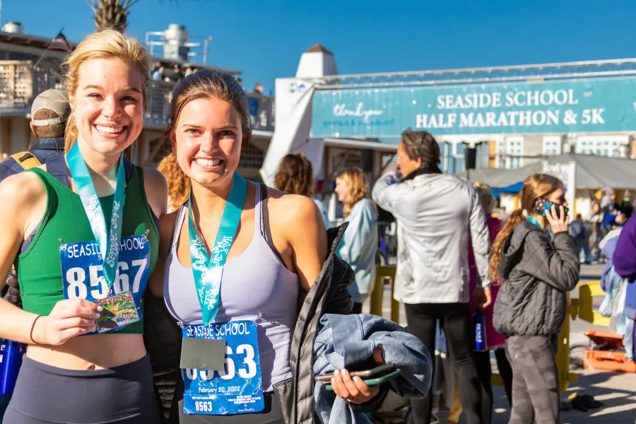 Two smiling women wearing race medals and bibs at the Seaside School Half Marathon & 5K.
