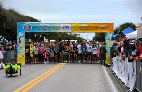 Runners lined up at the start line of a race, with a banner overhead and spectators on the side.