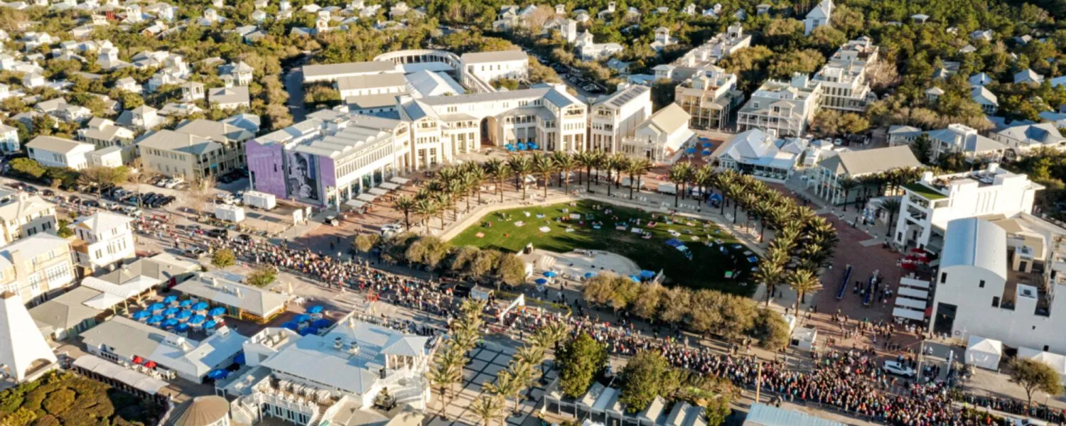 Aerial view of a large crowd of runners gathered on a street lined with buildings and palm trees, with people picnicking on a grassy area in the background.