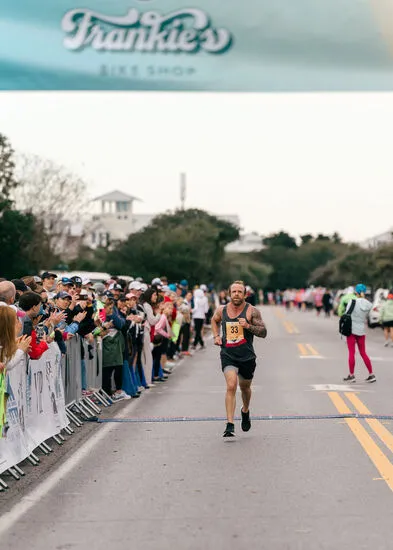 A male runner with tattoos crosses the finish line of a race, with a cheering crowd on the left and a sponsor banner overhead.
