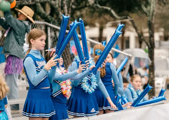 Young people in blue uniforms and leis holding blue inflatable noisemakers, cheering at an event.