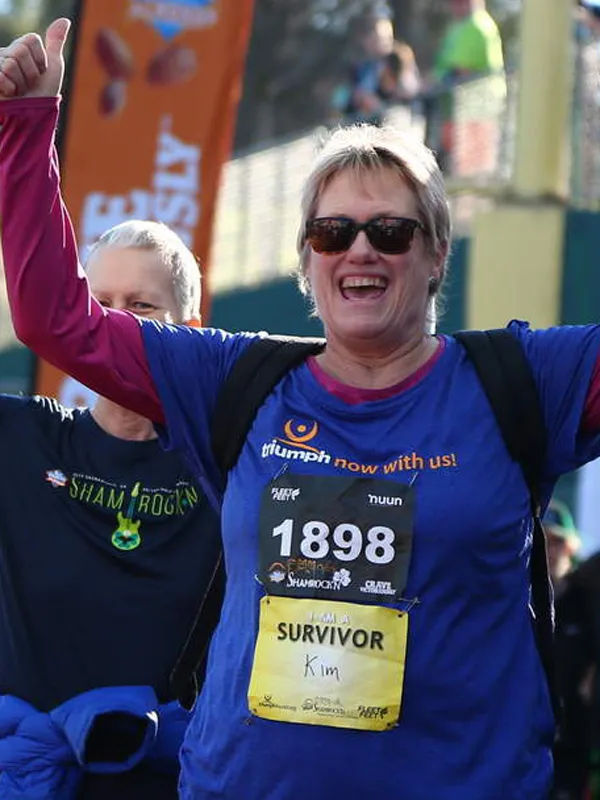 A woman with sunglasses and a blue shirt with a bib that says "1898 SURVIVOR Kim" smiles and gives a thumbs up, celebrating at a race.