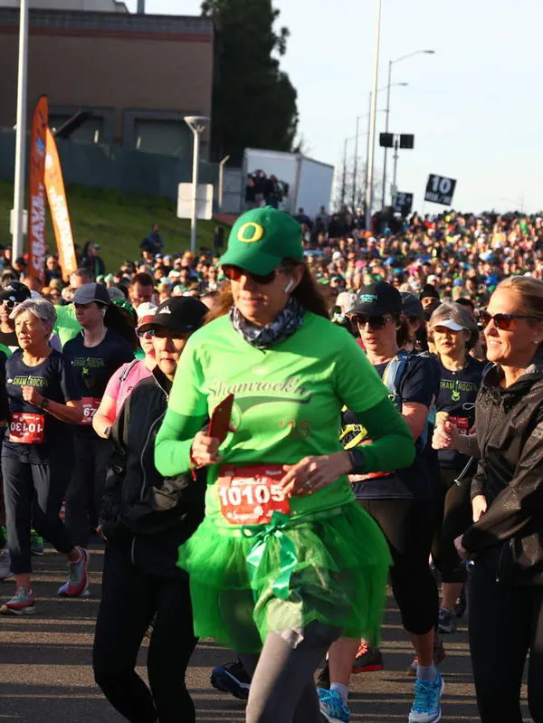 A group of runners, many wearing green, participate in the Shamrock 'n' Half Marathon. One runner in the foreground wears a green tutu and a shirt that says 'Shamrock's'.