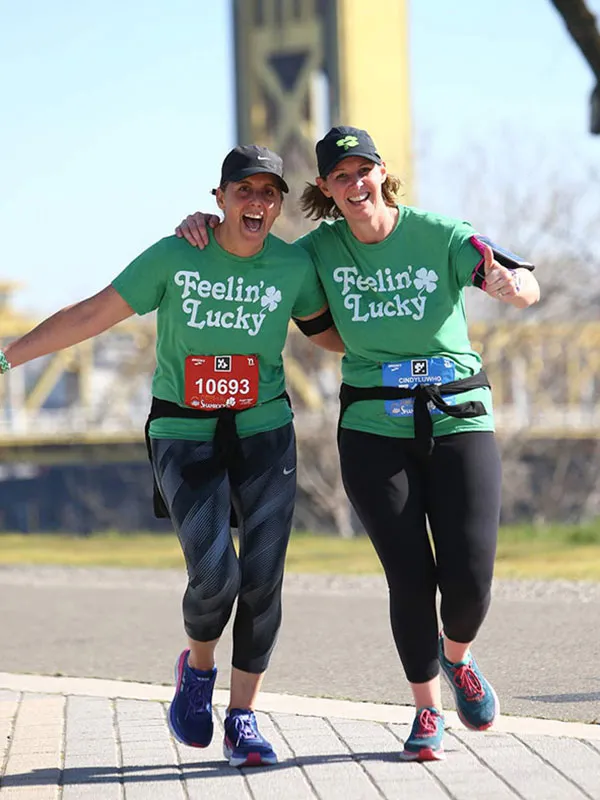 Two women in green 'Feelin' Lucky' shirts run together, smiling and looking excited, with one giving a thumbs-up.