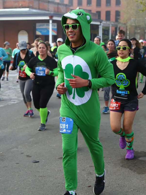A runner in a green costume with a shamrock design runs alongside other participants in a race.