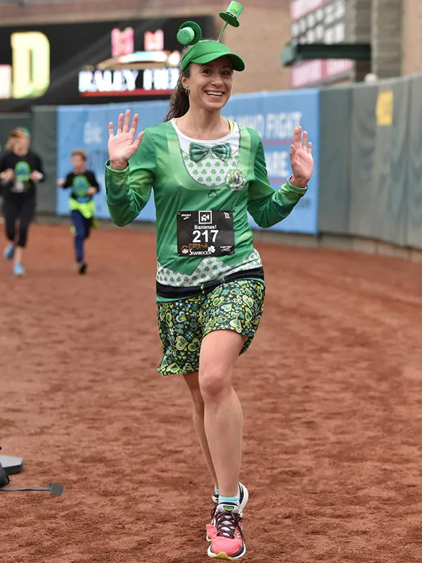 A woman in a green leprechaun costume with a shamrock hat runs and waves at the camera during a race.