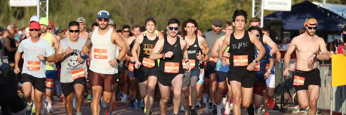 A group of runners in a race, with several in the foreground wearing bibs and running towards the camera.