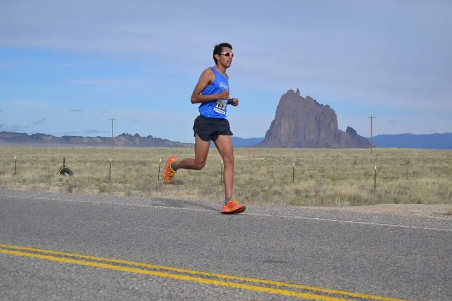 A male runner in a blue tank top and black shorts runs on a paved road with yellow lines, with a distinctive rock formation in the background under a clear blue sky.