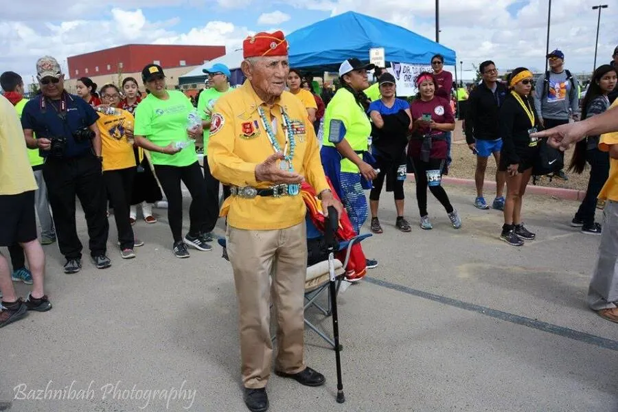 An elder in traditional attire stands with a cane, surrounded by runners and spectators before a race.