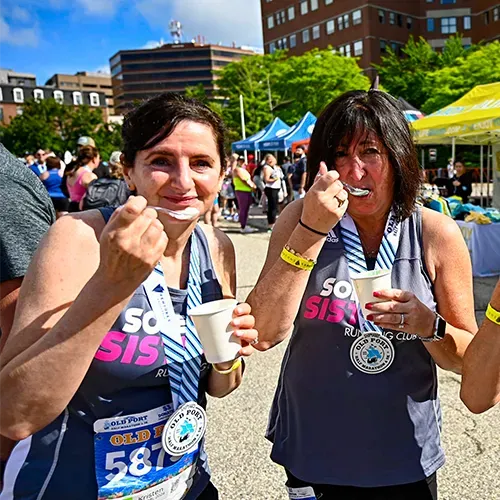 Two women wearing race medals and bibs eat ice cream after a race.