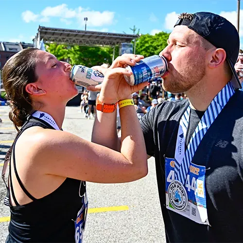 A man and a woman wearing race medals drink from cans after completing a race.