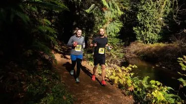 Two runners smiling as they run on a wooded trail next to a stream.