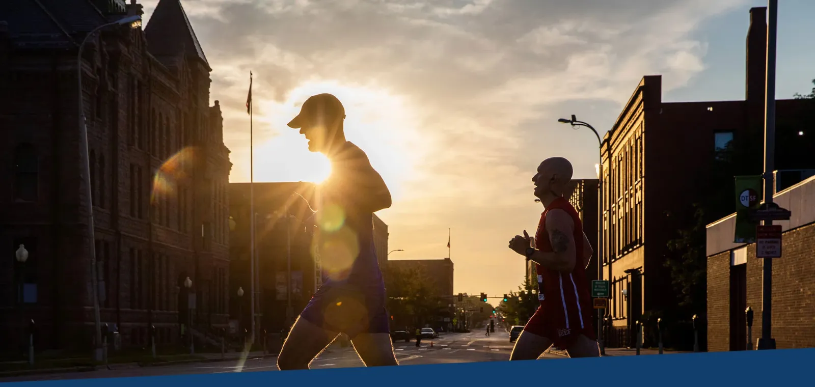Two runners silhouetted against a sunset on an urban street, with historic buildings on either side.