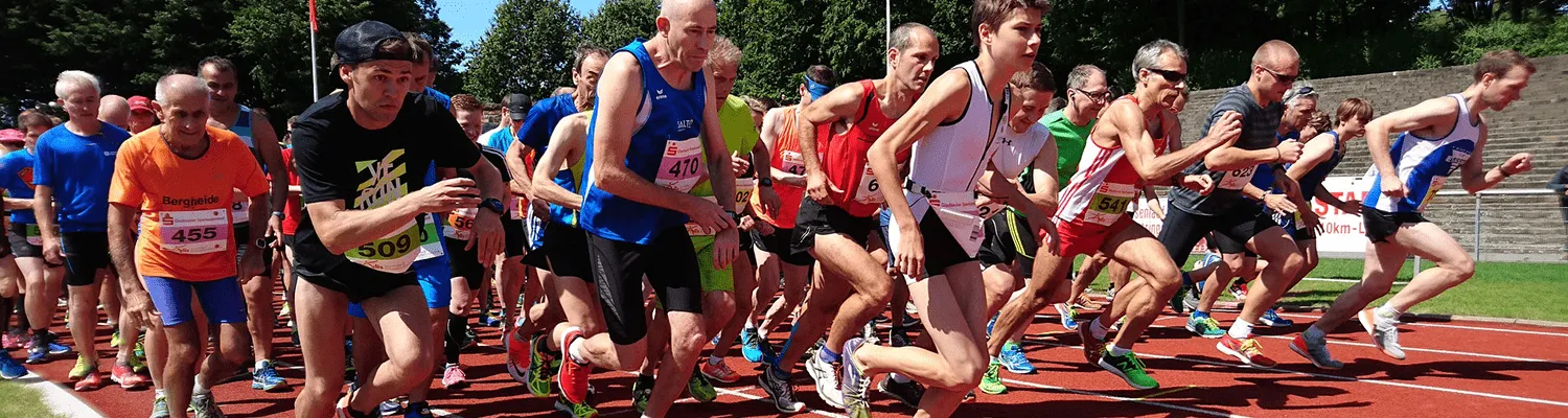 A group of runners in athletic wear are lined up on a track, some with bib numbers visible, appearing to be at the start of a race.