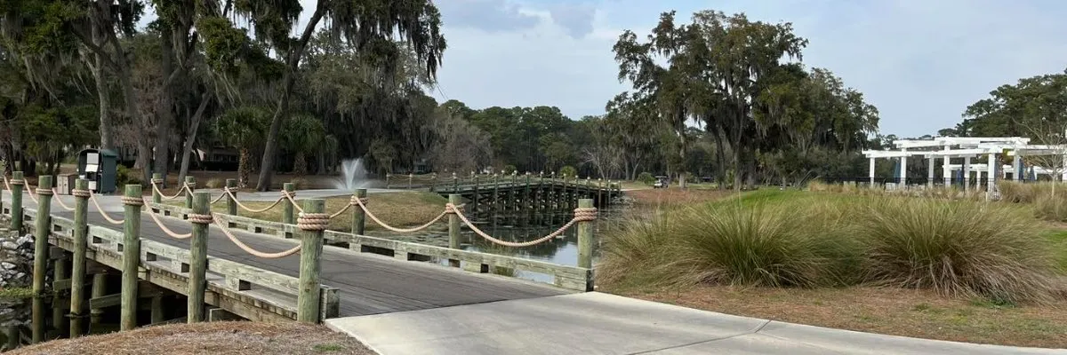 A wooden bridge with rope railings crosses over water, with trees draped in Spanish moss and a white pergola structure in the background.
