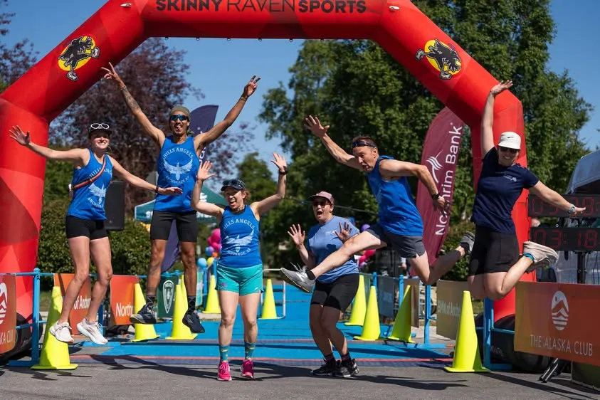 A group of runners in blue shirts jump in the air with excitement under a red finish line arch at the Skinny Raven Sports race.