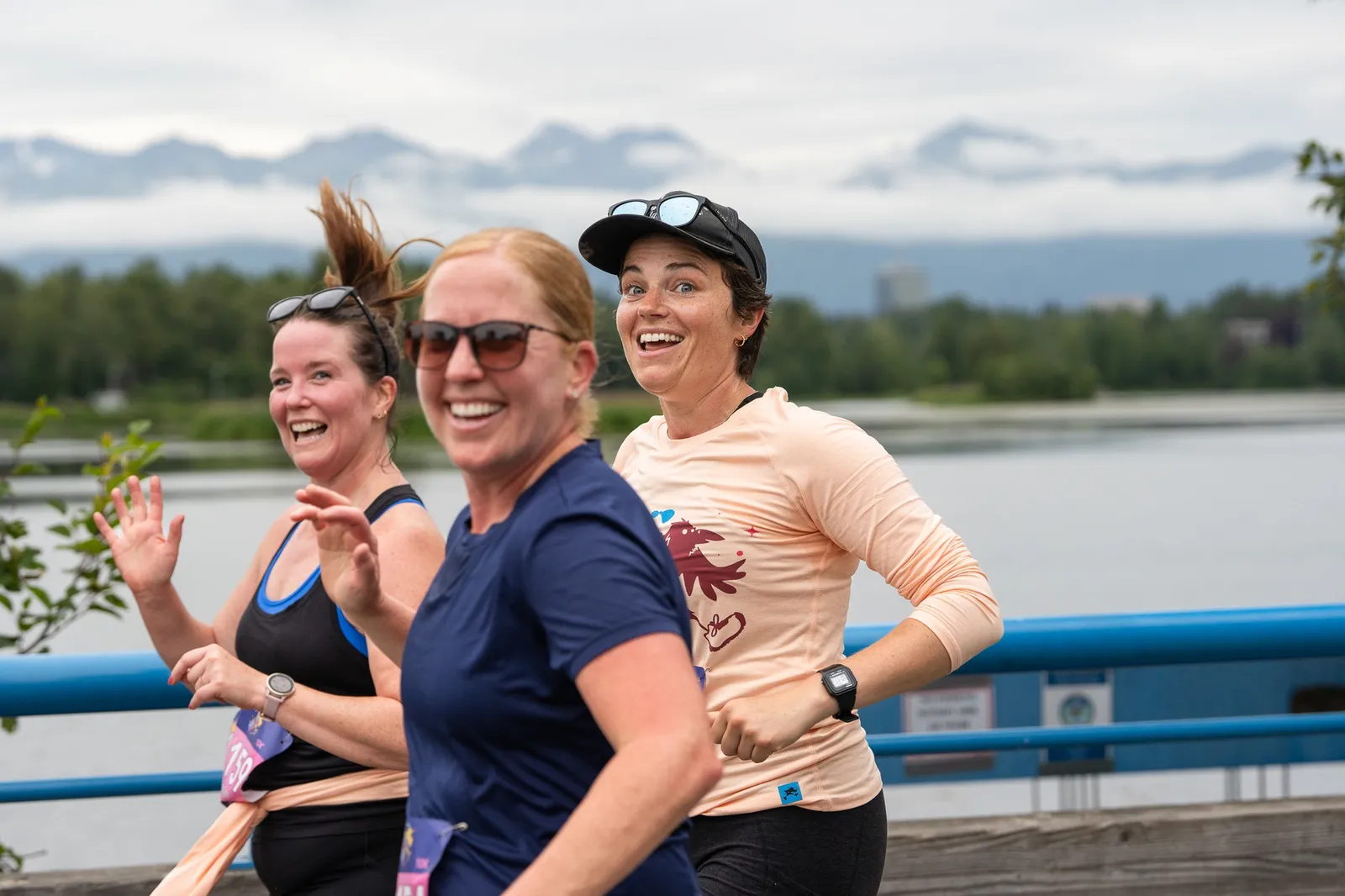 Three women smiling and running along a waterfront path with mountains in the background.
